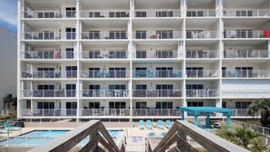 A multi-story hotel with balconies, a pool area with lounge chairs, and a wooden deck leading to the pool.