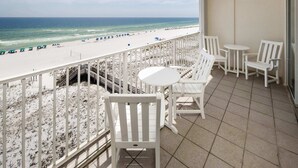 A balcony with white chairs and a small table, overlooking a beach with umbrellas and people.