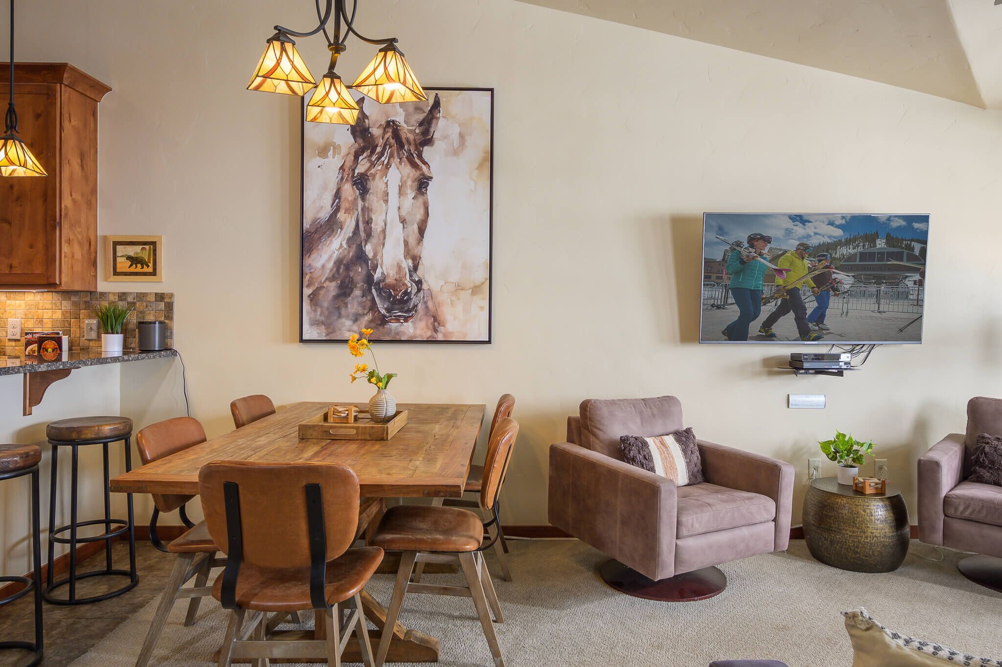 Un coin repas avec une table en bois, des chaises et des luminaires suspendus. Un grand tableau orne le mur et un téléviseur est fixé au-dessus de la table.