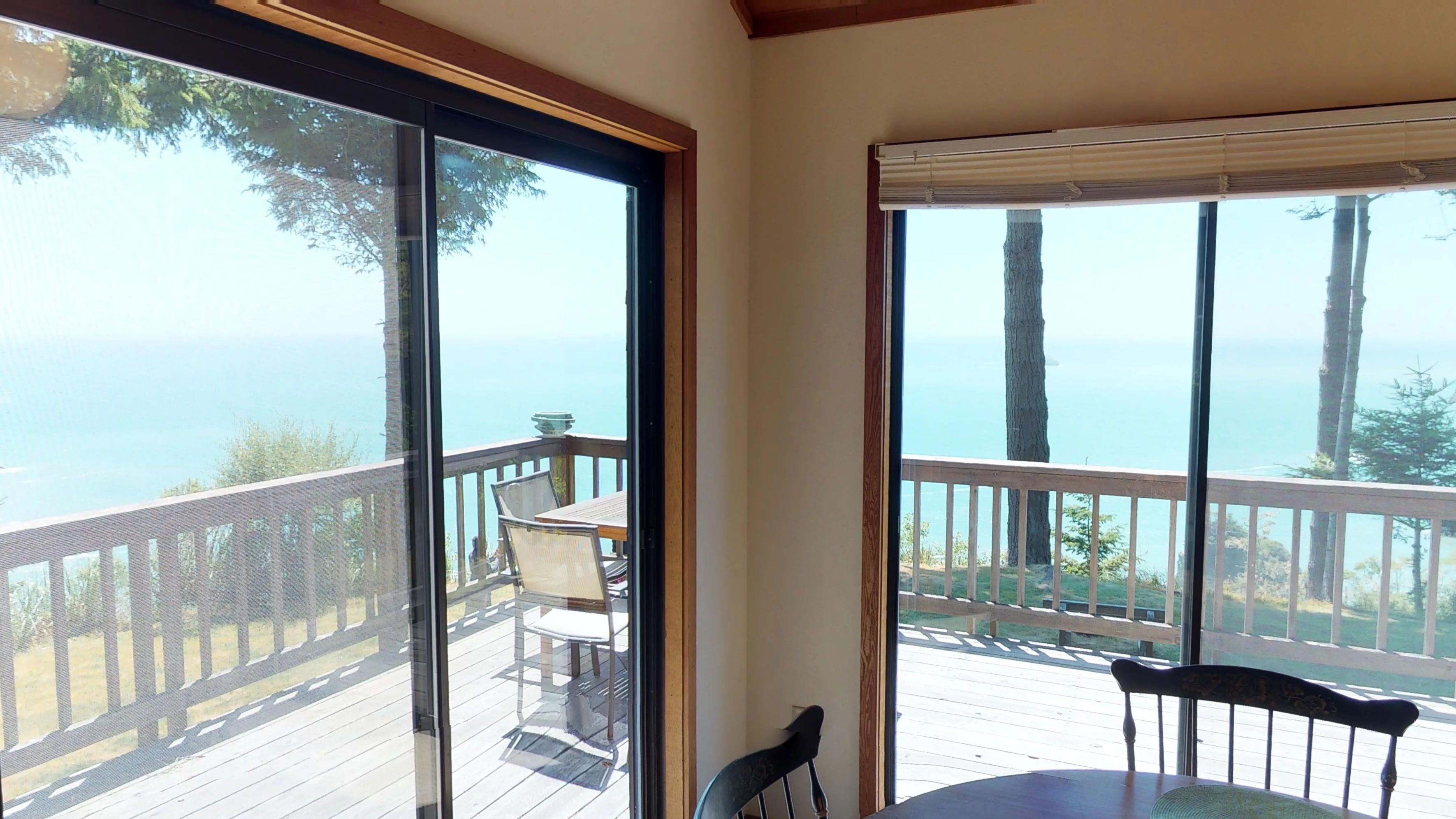 A room with large windows offering a view of the ocean. There is a wooden deck outside with a table and chairs.