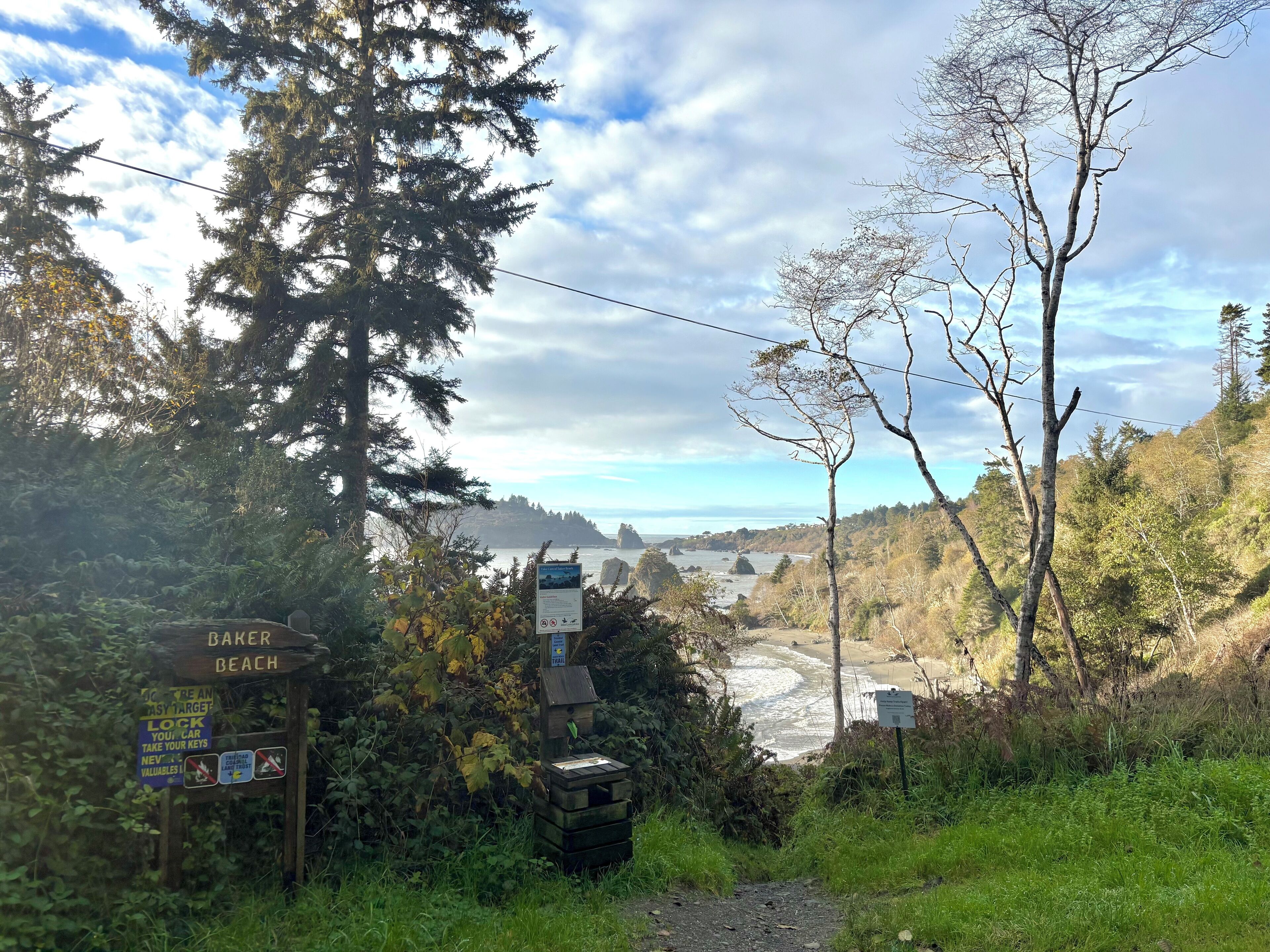 Ein Wegweiser mit der Aufschrift 'Baker Beach' und verschiedenen Hinweisschildern, umgeben von üppiger Vegetation und Blick auf den Strand und das Meer.