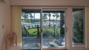 A room with a sliding glass door leading to a pool area with a view of palm trees and a clear sky. - 1865 Beachside Tennis- Easy Access to Beach & Pool. (Hilton Head Island)