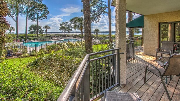 A pool area with a view of the ocean, surrounded by palm trees and a wooden deck with chairs. - 1865 Beachside Tennis- Easy Access to Beach & Pool. (Hilton Head Island)