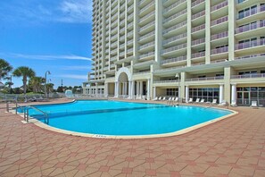 Une grande piscine extérieure dotée d’une terrasse hexagonale carrelée, entourée d’un hôtel à plusieurs étages avec des balcons et d’un espace couvert aménagé avec des chaises longues.
