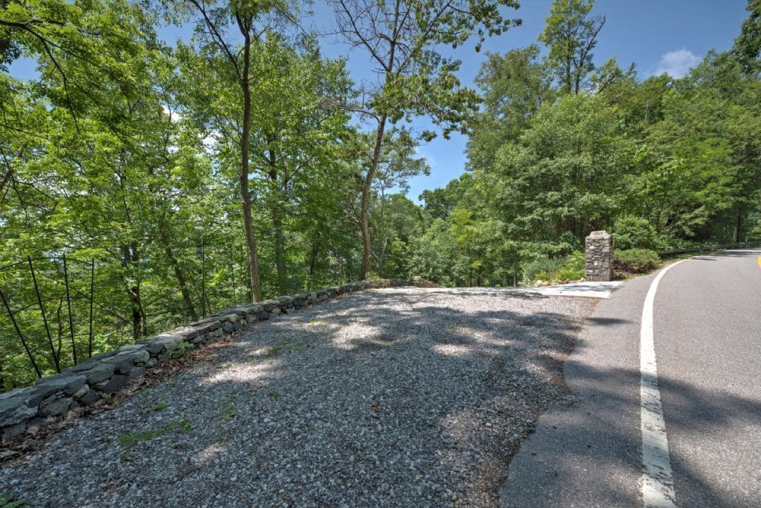 A gravel path with a stone wall on one side and a road on the other, surrounded by dense green trees.