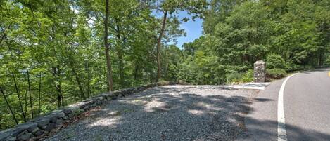A gravel path with a stone wall on one side and a road on the other, surrounded by dense green trees.