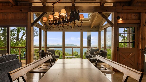 Interior de madera con una gran mesa y bancos, ventanas amplias con vistas a montañas, y un candelabro.