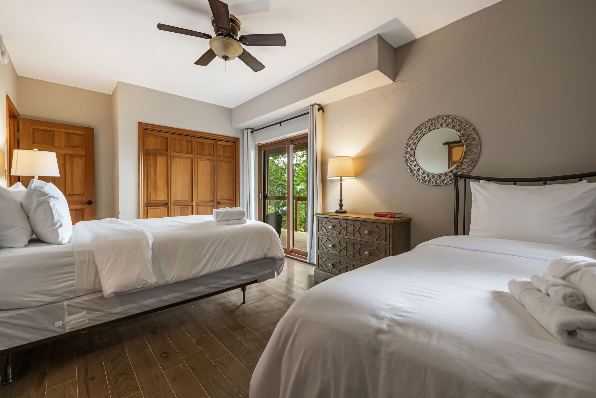 A hotel room with two beds, a ceiling fan, a wooden wardrobe, a dresser, and a large window with a view of greenery.