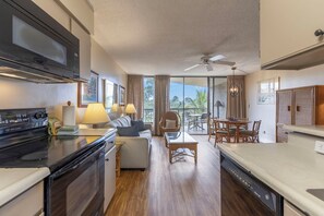 A modern kitchen with a stove, oven, and dishwasher. There is a dining area with a table and chairs, and a sofa. A ceiling fan is visible. - Stunning views from condo suite featuring private lanai deck (Kihei)
