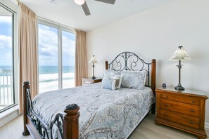 Une chambre avec un grand lit, deux tables de chevet avec des lampes, une commode en bois et une vue sur l’océan grâce à de grandes fenêtres.