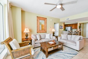 A living room with a sofa, a wooden coffee table, and a wicker chair. There is a ceiling fan and a kitchen in the background.