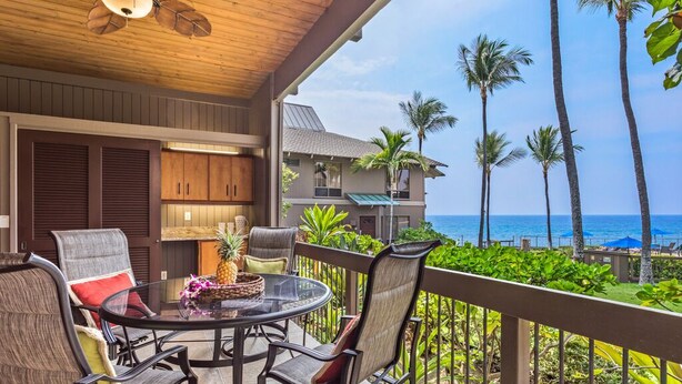 A covered outdoor area with a table and chairs, a ceiling fan, and a view of the ocean with palm trees. - Aloha Condos, Kanaloa at Kona, Condo 2602, Oceanfront, AC (Kailua Kona)