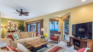 A living room with a sofa, coffee table, and TV, a ceiling fan, and a view of the ocean. - Aloha Condos, Kanaloa at Kona, Condo 2602, Oceanfront, AC (Kailua Kona)