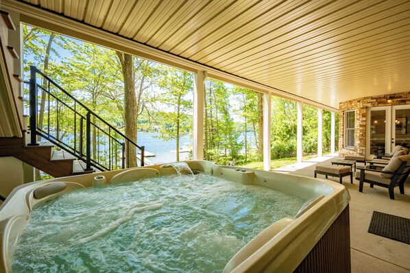 A hot tub with a view of a lake, a covered patio with outdoor furniture, and a staircase leading to an upper level.