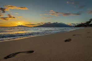 A beach with footprints leading to the water, a sunset, and a mountain in the background.
