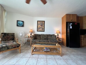 A living room with a sofa, armchair, and coffee table, a refrigerator, and a fan.