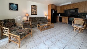 Una sala de estar con un sofá, dos sillones, una mesa de centro, un ventilador y un ventilador de techo. Hay una zona de cocina con armarios de madera y electrodomésticos.