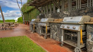 Una hilera de parrillas para barbacoa con un fondo de pared de piedra y un área de estar cubierta con mesas de picnic.