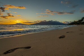 Una spiaggia con impronte che conducono all'acqua, un tramonto e una montagna sullo sfondo.