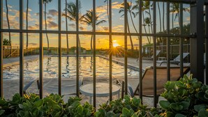 Zona de piscina con vista al atardecer, rodeada de palmeras y una cerca metálica.
