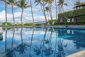 A pool with clear water reflecting palm trees, surrounded by lounge chairs and a fence.