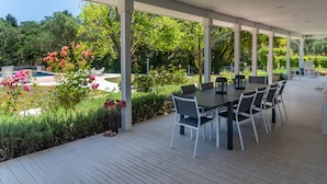 An outdoor patio with a table and chairs, surrounded by lush greenery and a swimming pool.