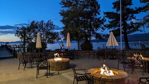 An outdoor patio with stone fire pits and tables, overlooking a body of water and mountains.