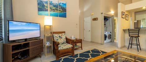 A living room with a television, a sofa, a coffee table, and a rug. There is a door leading to a laundry area.