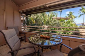 A balcony with a round table and two chairs, overlooking a tropical garden with palm trees and a view of the ocean.
