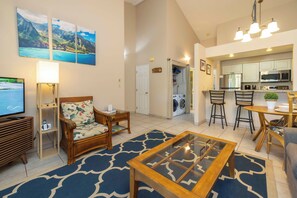 A living room with a sofa, coffee table, and a TV. There is a kitchen area with a dining table and chairs.