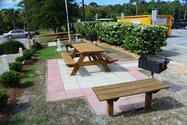 Mesas de picnic al aire libre con un contenedor de basura, un seto y un edificio amarillo al fondo.