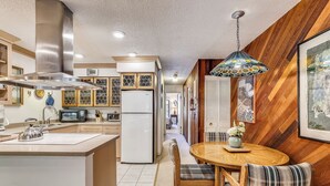 A modern kitchen with a central island, stainless steel range hood, and a dining area with a round wooden table and plaid chairs. - Charming condo in the core, dynamite location (Aspen)