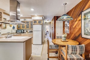 A modern kitchen with a central island, stainless steel range hood, and a dining area with a round wooden table and plaid chairs. - Charming condo in the core, dynamite location (Aspen)