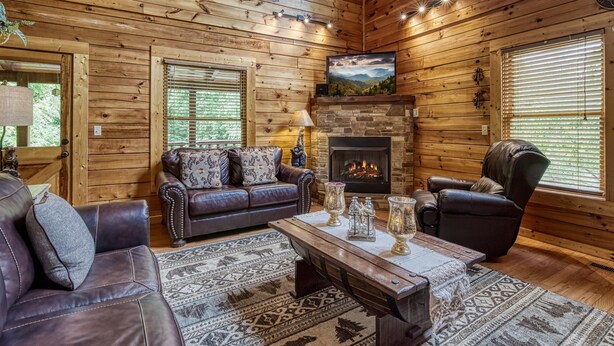 A cozy log cabin interior with a fireplace, leather sofas, and a large window.