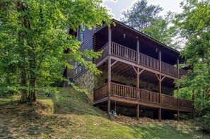 A two-story wooden cabin with a stone foundation, surrounded by lush greenery.