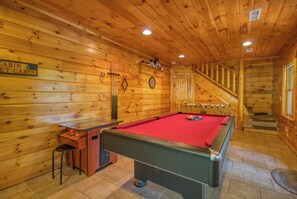 Interior de una cabaña de madera con mesa de billar, dardos y una escalera que lleva a una zona tipo altillo.