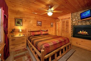 Un dormitorio rústico de cabaña de madera con cama, mesitas de noche, lámpara, chimenea y televisor.