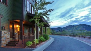 A two-story building with a stone facade, a covered porch, and a view of mountains in the background. - Cozy Luxury Studio with Fireplace, Prime Location & Summer Adventure! (Deer Valley)