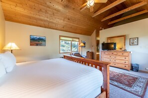 Een slaapkamer met een groot bed, houten balken in het plafond, een dressoir, een televisie en een raam met uitzicht op bomen.