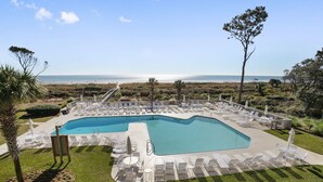 A pool area with lounge chairs and umbrellas, surrounded by greenery and a view of the ocean.