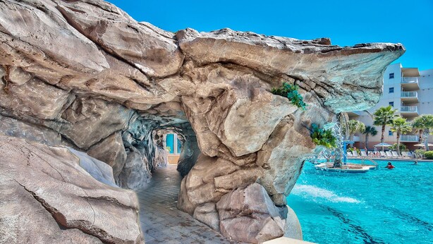 Formação rochosa com entrada em túnel, área de piscina com espreguiçadeiras e um edifício ao fundo.