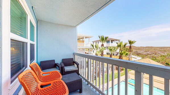 A balcony with wicker chairs and a small orange chair, overlooking a pool and beachfront properties.