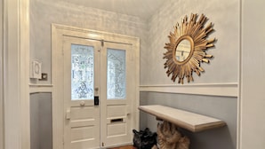 Un pasillo con una puerta blanca con paneles de vidrio esmerilado, un espejo decorativo y un estante pequeño.