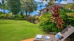 A well-maintained lawn with a lounge chair and a book, surrounded by tropical plants and a house with a view of the ocean. - Kahala 713 Poipu Beach Ocean By Suite Paradise (Koloa)