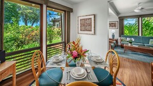 A dining area with a table set for four, large windows with a view of greenery, and a seating area with a sofa and a coffee table. - Kahala 713 Poipu Beach Ocean By Suite Paradise (Koloa)