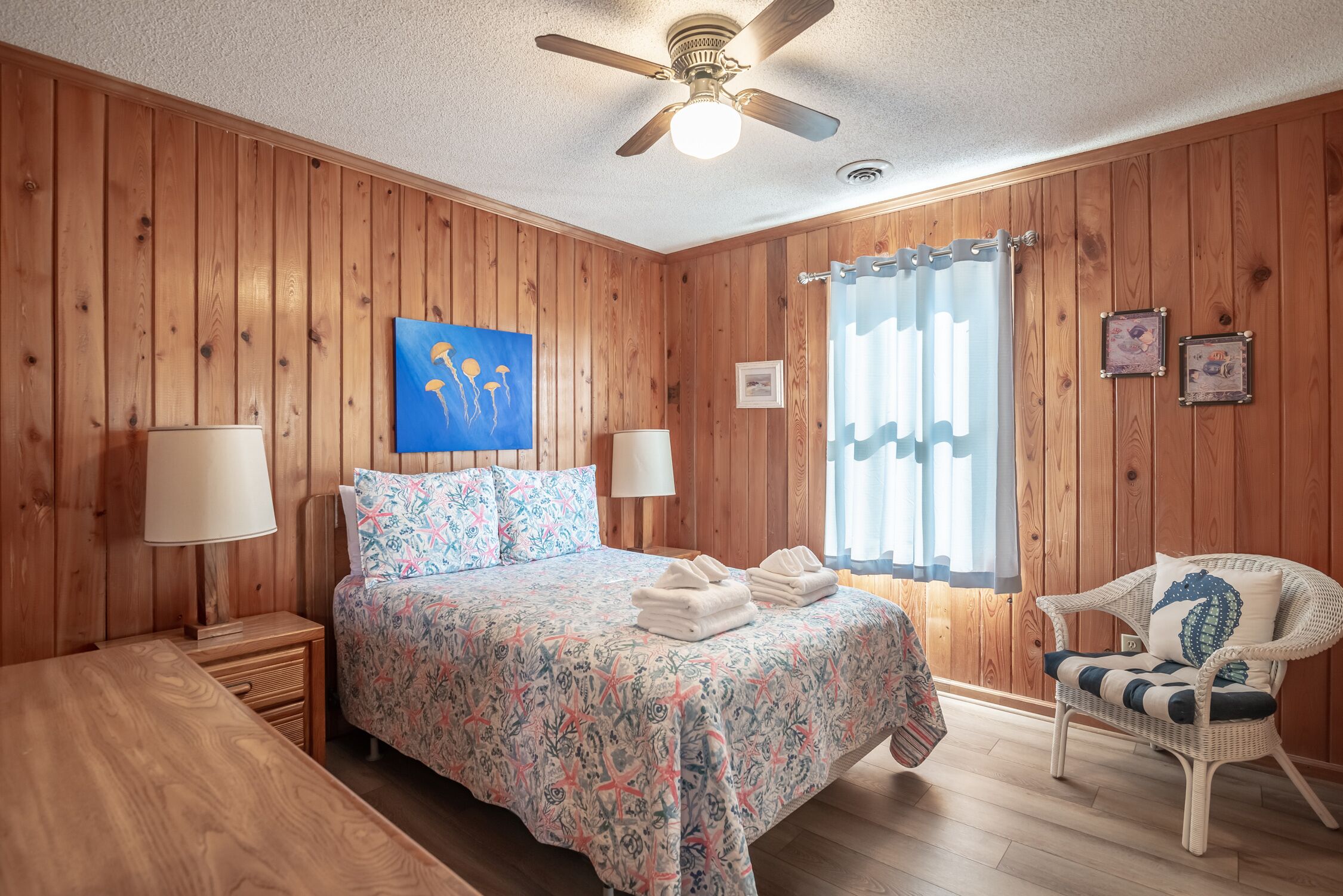 A wooden-paneled room with a bed, two bedside lamps, a ceiling fan, a window with curtains, and a wicker chair.