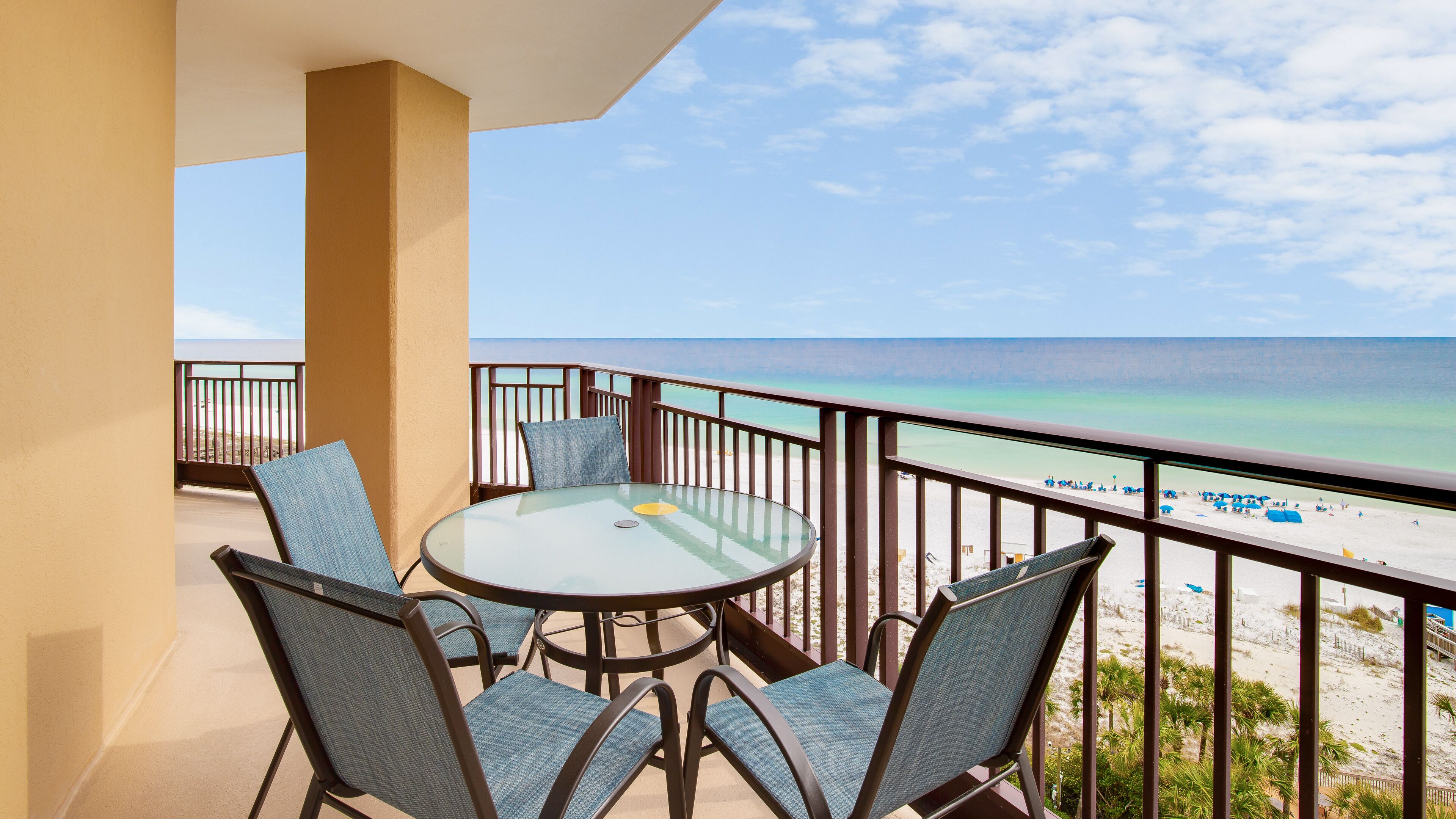 A balcony with a glass table and four chairs, overlooking a beach with clear turquoise water and a sandy shore.