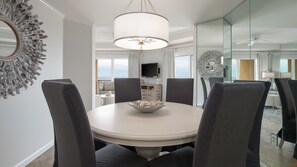 A dining area with a round table, grey upholstered chairs, a central chandelier, and a large mirror on the wall.