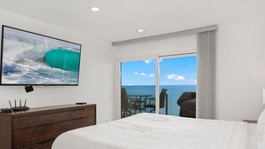 A hotel room with a large bed, a TV mounted on the wall, a wooden dresser, and a sliding glass door leading to a balcony with a view of the ocean.