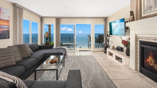 A spacious living room with a large sofa, a coffee table, and a fireplace. There are multiple windows offering a view of the ocean.